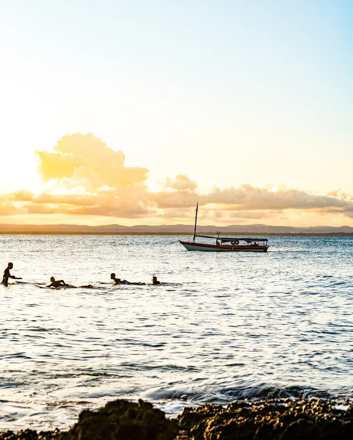 Banhistas e um barco ao pôr do sol, com recifes em primeiro plano
